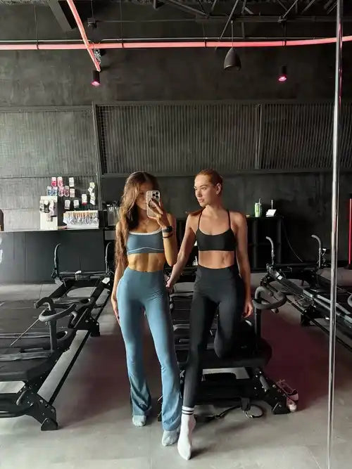 Two women posing in a gym setting, wearing matching athletic wear. One is wearing a light blue sports bra and matching flared leggings, while the other wears a black sports bra and black leggings.